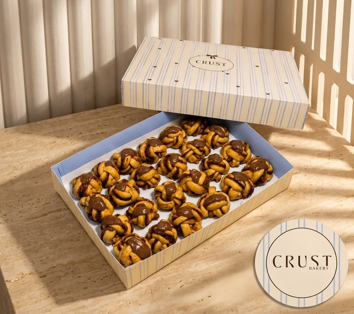 An open CRUST box, cream with soft blue stripes, filled with rows of hand tied knots under a chocolate drizzle.